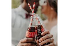 Two people holding coca-cola bottles cheersing with paper straws inside the bottles.
